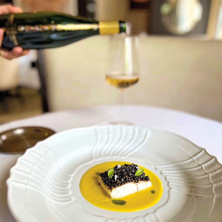 A member of Fiola's staff pouring sparkling wine into a wineglass on a table near a plate of Mediterranean sea bass and lentils